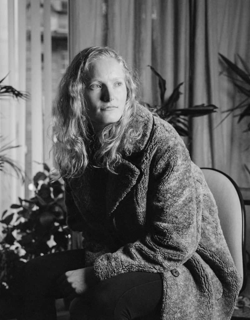 Black and white portrait of Rachel Byrne, sitting on a chair in a dark studio.